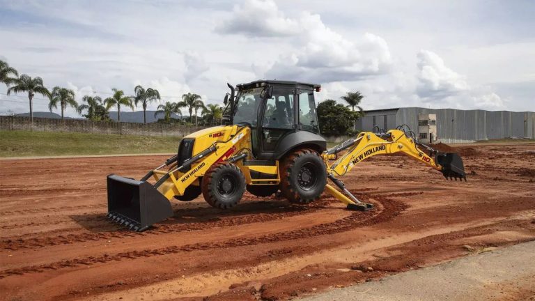 Retroescavadeira amarela da marca New Holland estacionada em um terreno de terra, com a caçamba frontal e o braço de escavação traseiro apoiados no solo sob um céu nublado.