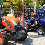 Fileira de tratores compactos de diferentes marcas expostos lado a lado em feira agrícola, com destaque para um modelo laranja em primeiro plano.