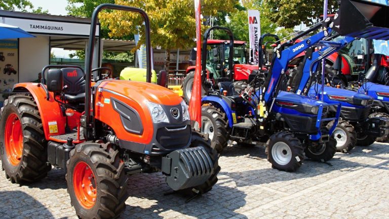 Fileira de tratores compactos de diferentes marcas expostos lado a lado em feira agrícola, com destaque para um modelo laranja em primeiro plano.
